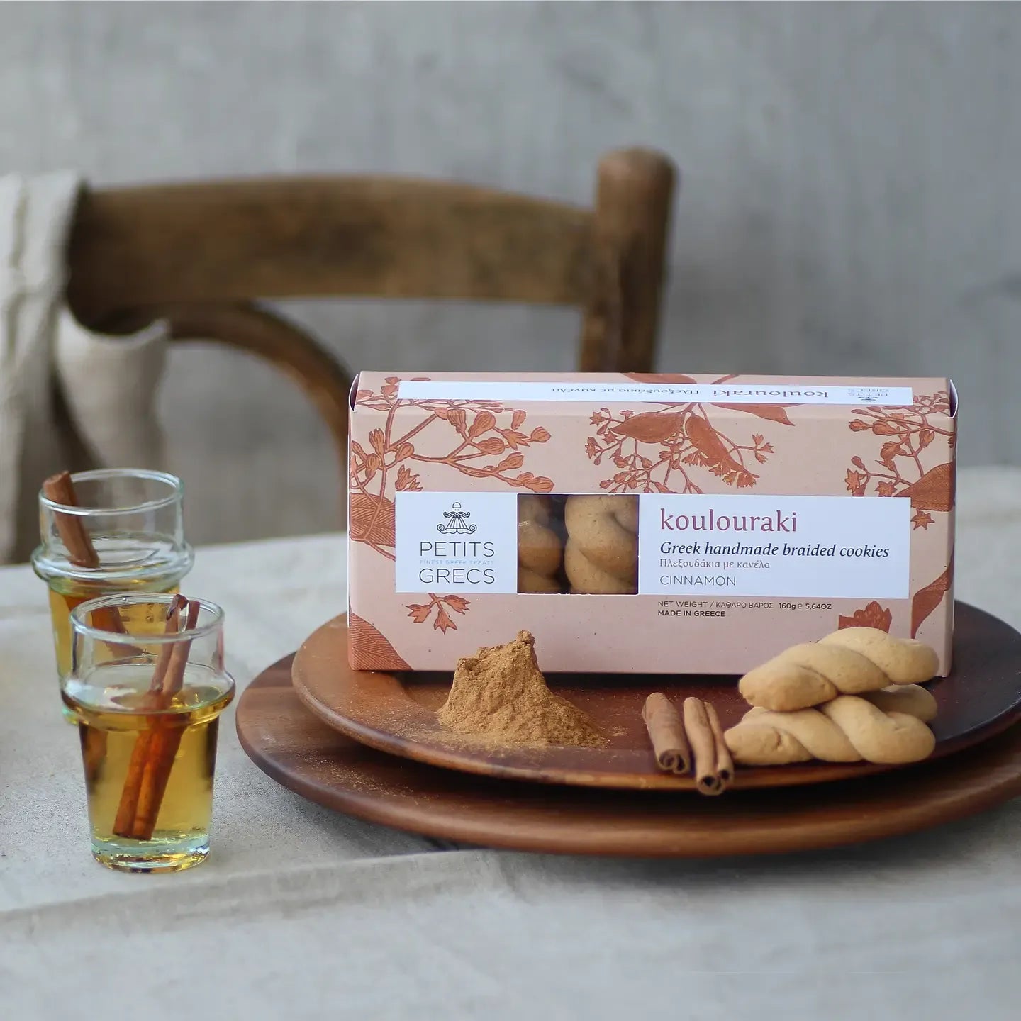 Box of koulouraki cookies with a plate of cookies, cinnamon sticks, and a glass of honey on a table