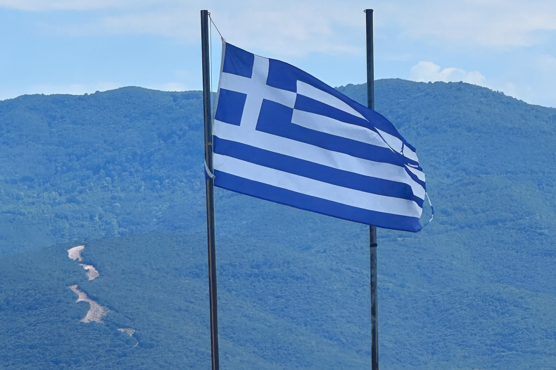 Greek flag waiving proudly from Nea Vrasna Beach in front of mountains of Stavros Greece