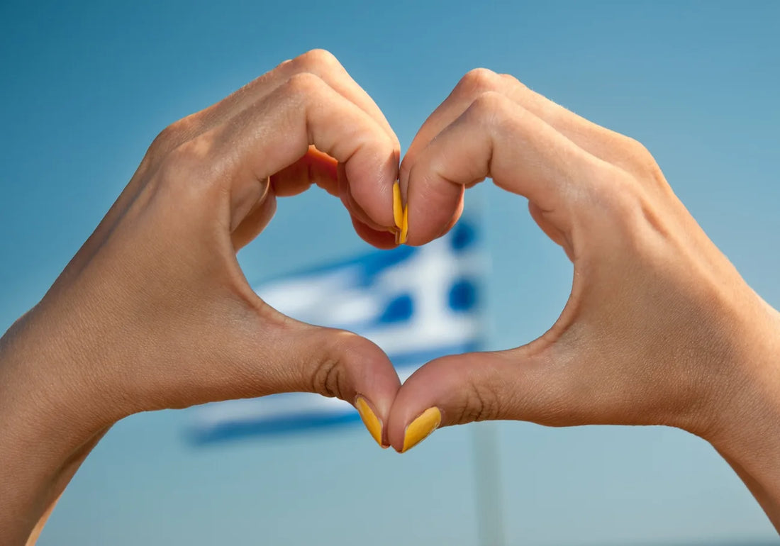 Heart hands with yellow nail polish in front of Greek flag and blue sky