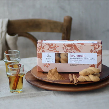 Box of koulouraki cookies with a plate of cookies, cinnamon sticks, and a glass of honey on a table
