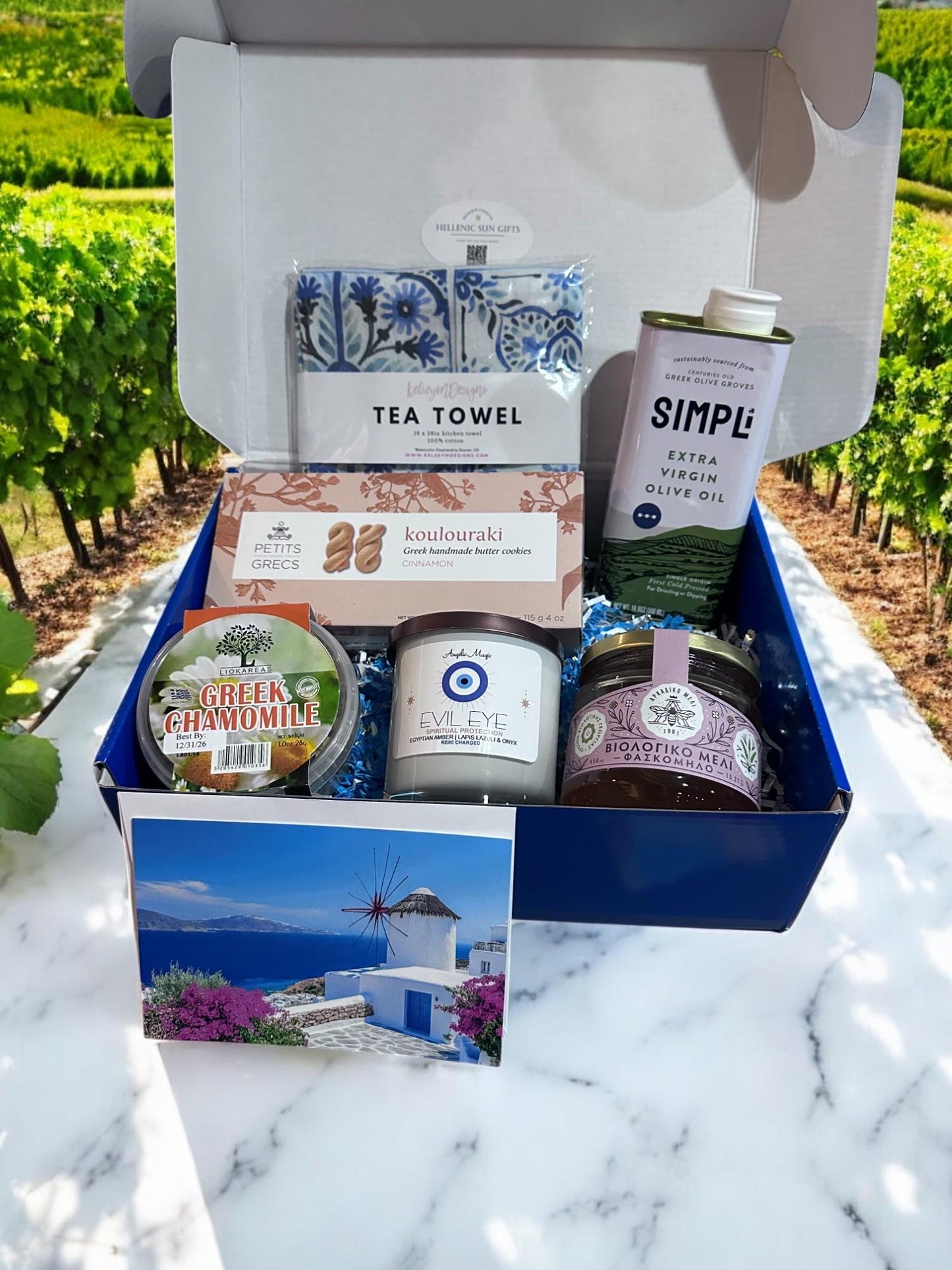 Gift box with Greek-themed products including olive oil, tea towel, and candles, set against a vineyard background.