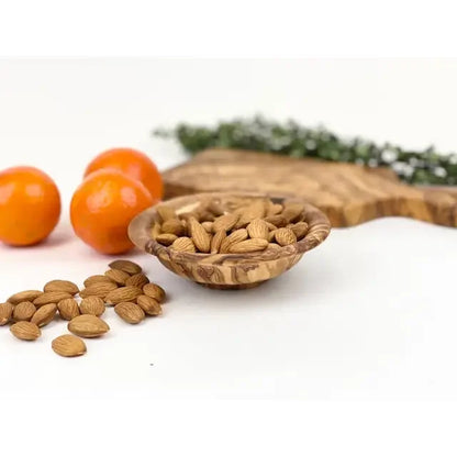 Wooden bowl filled with almonds on a white surface with oranges and greenery in the background