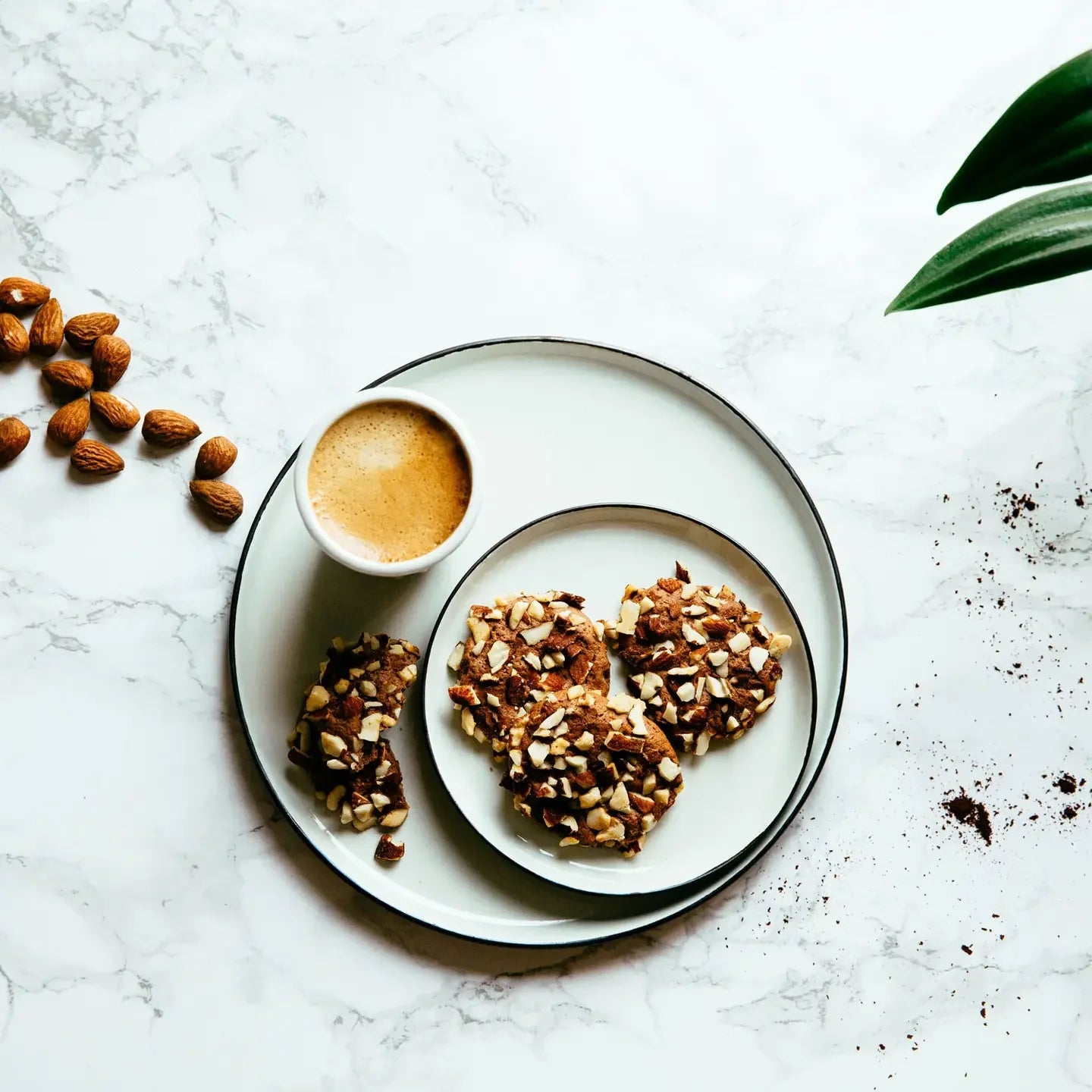 Three cocoa almond cookies on a plate with a small cup of coffee and scattered almonds on a marble surface.