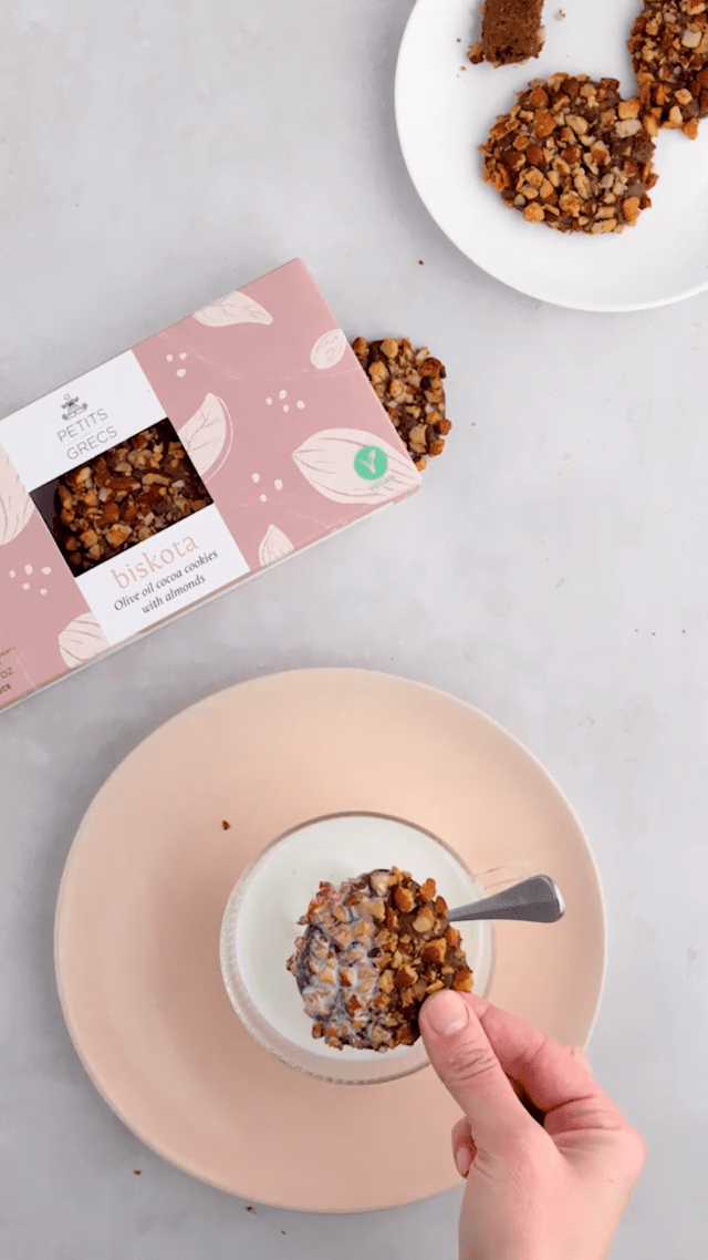 Person dipping cocoa almond cookie into a glass of milk with a spoon and a box of cocoa almond cookies and additional plate of cookies on a plate in the background