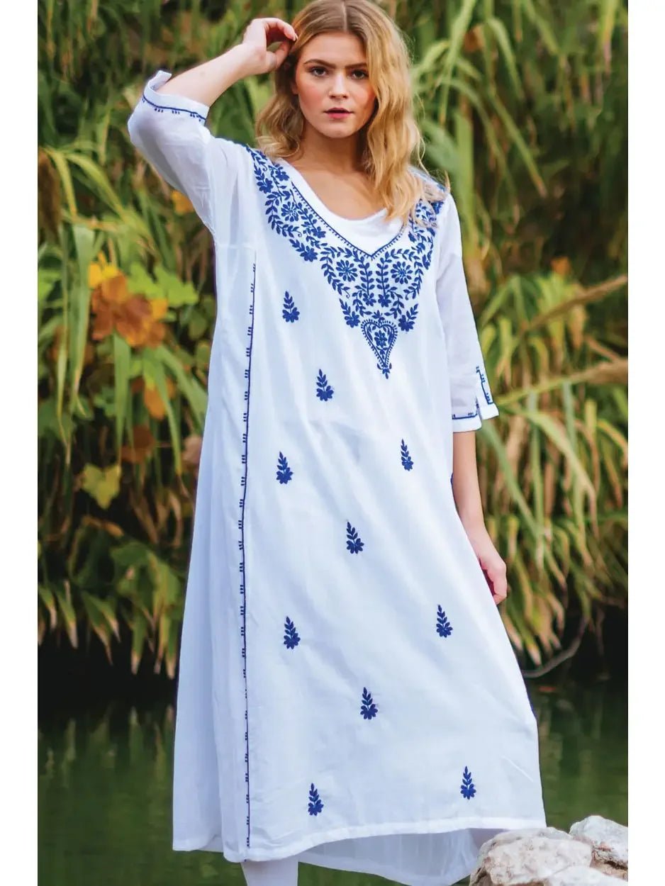 Woman wearing a white dress with blue embroidery standing outdoors near water and greenery.