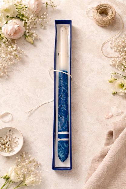 Candle in a blue decorative holder surrounded by flowers on a light surface