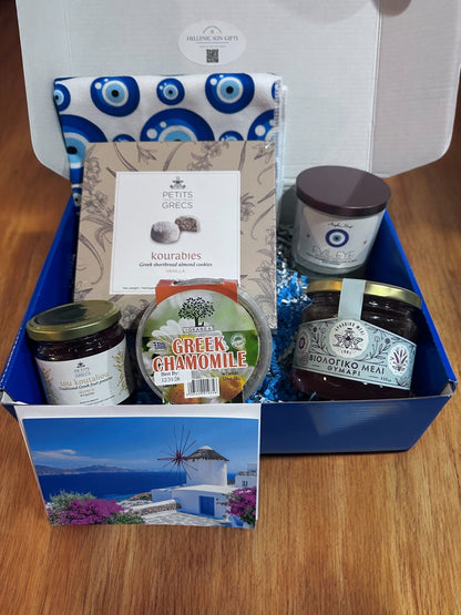 Gourmet gift box with Greek-themed products including tea towel, honey, candle, preserves, cookies and tea on a wooden surface