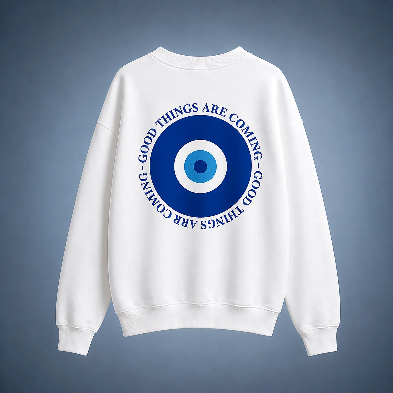 A white crewneck sweatshirt shown from the back, featuring a large blue and white evil eye graphic with the text 'GOOD THINGS ARE COMING' in blue around the eye design, set against a dark blue gradient background.