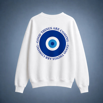 A white crewneck sweatshirt shown from the back, featuring a large blue and white evil eye graphic with the text 'GOOD THINGS ARE COMING' in blue around the eye design, set against a dark blue gradient background.