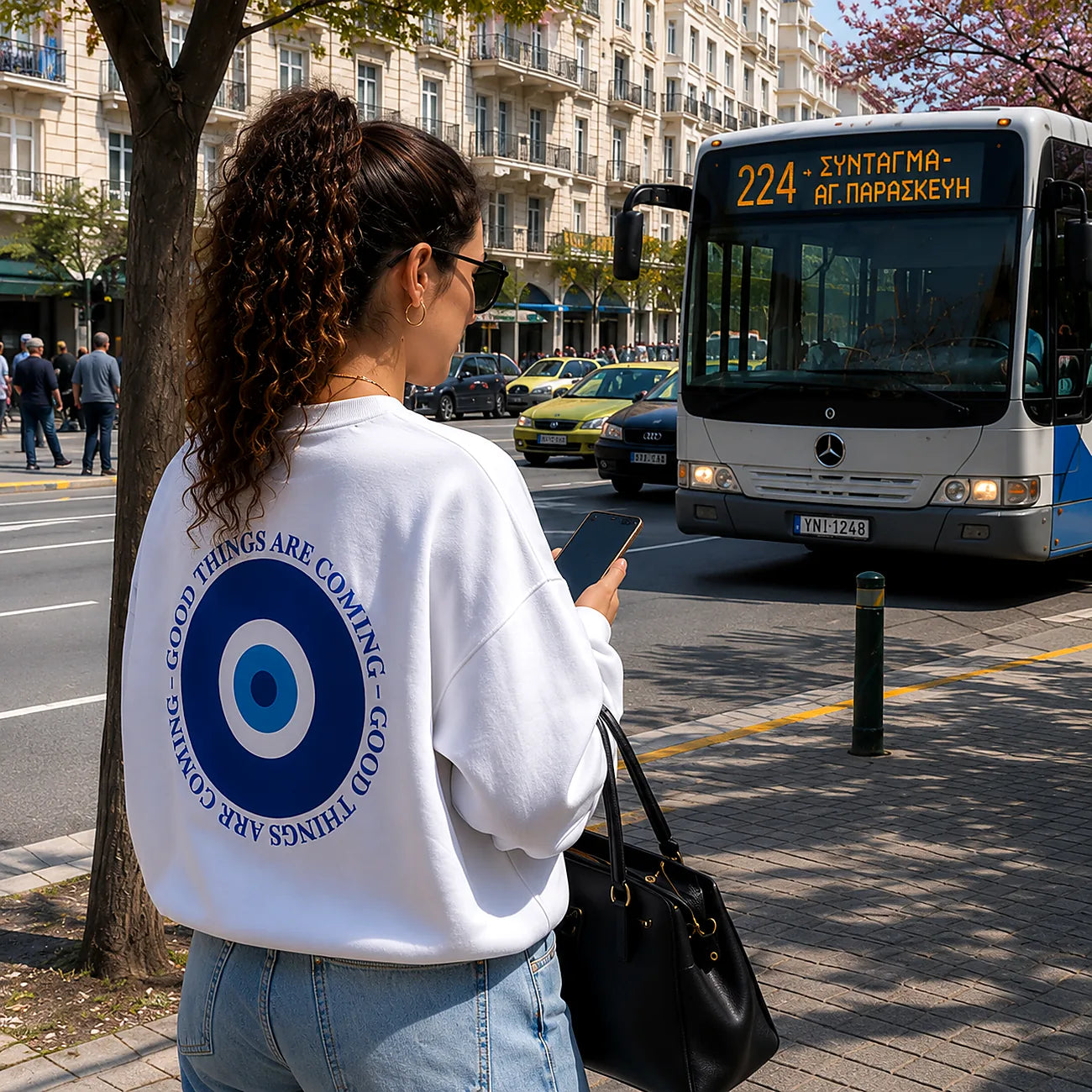 A woman stands on a city sidewalk facing a bus, wearing a white sweatshirt with a large blue circular graphic and text on the back, light blue jeans, and carrying a black handbag. The background shows a street scene with buildings, cars, and a bus labeled 224.