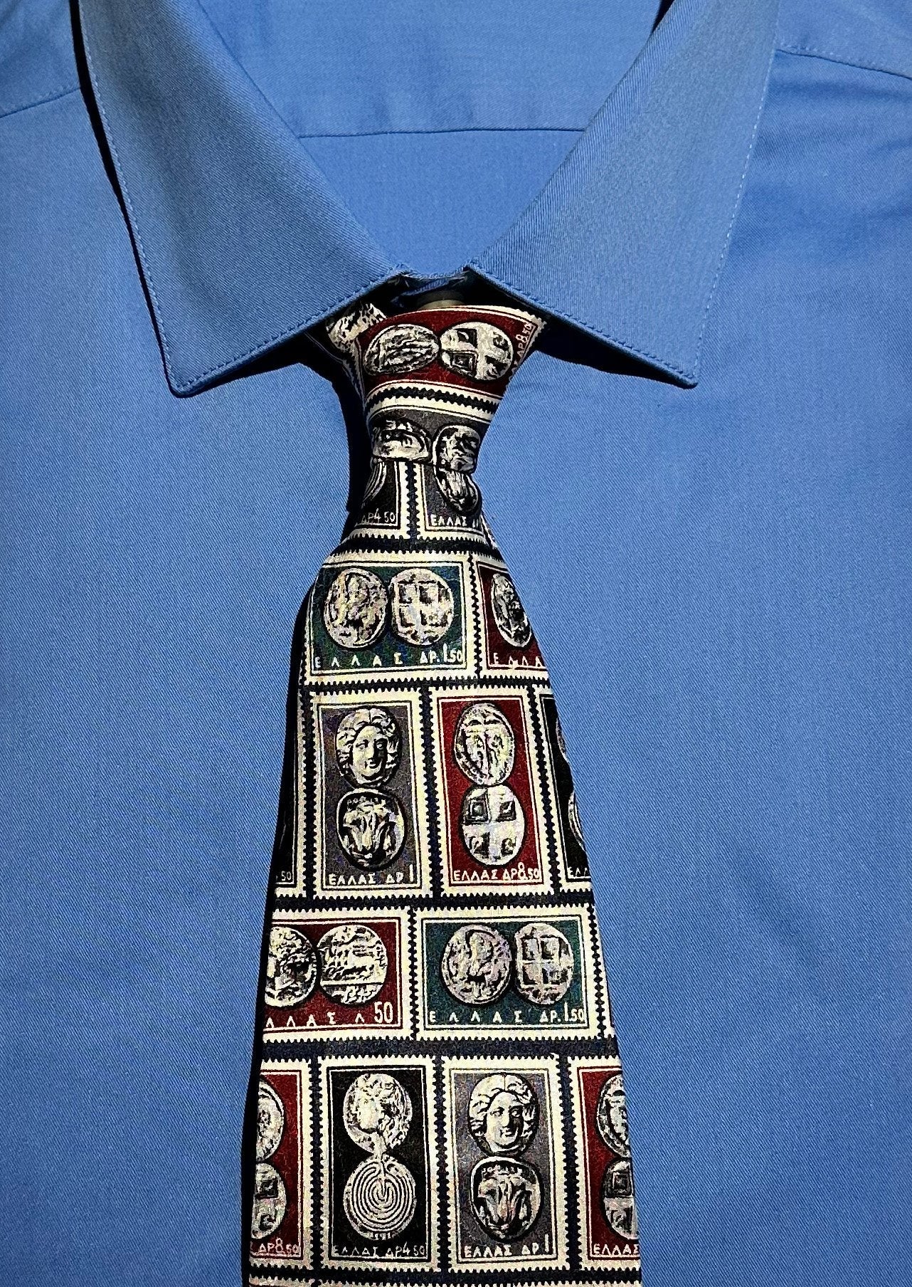 Close-up of a tie with a pattern of Greek stamps with ancient Greek coins in rectangles worn with a blue shirt