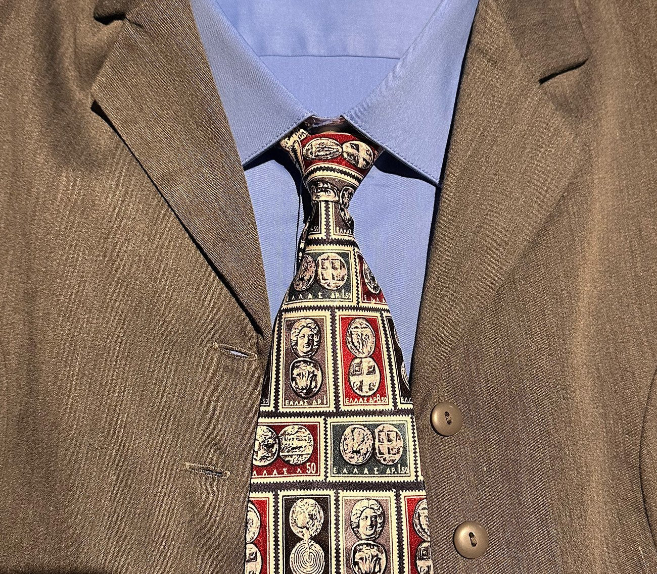 Close-up of a tie with a pattern of Greek stamps with ancient Greek coins in rectangles worn with a graysuit jacket and blue shirt