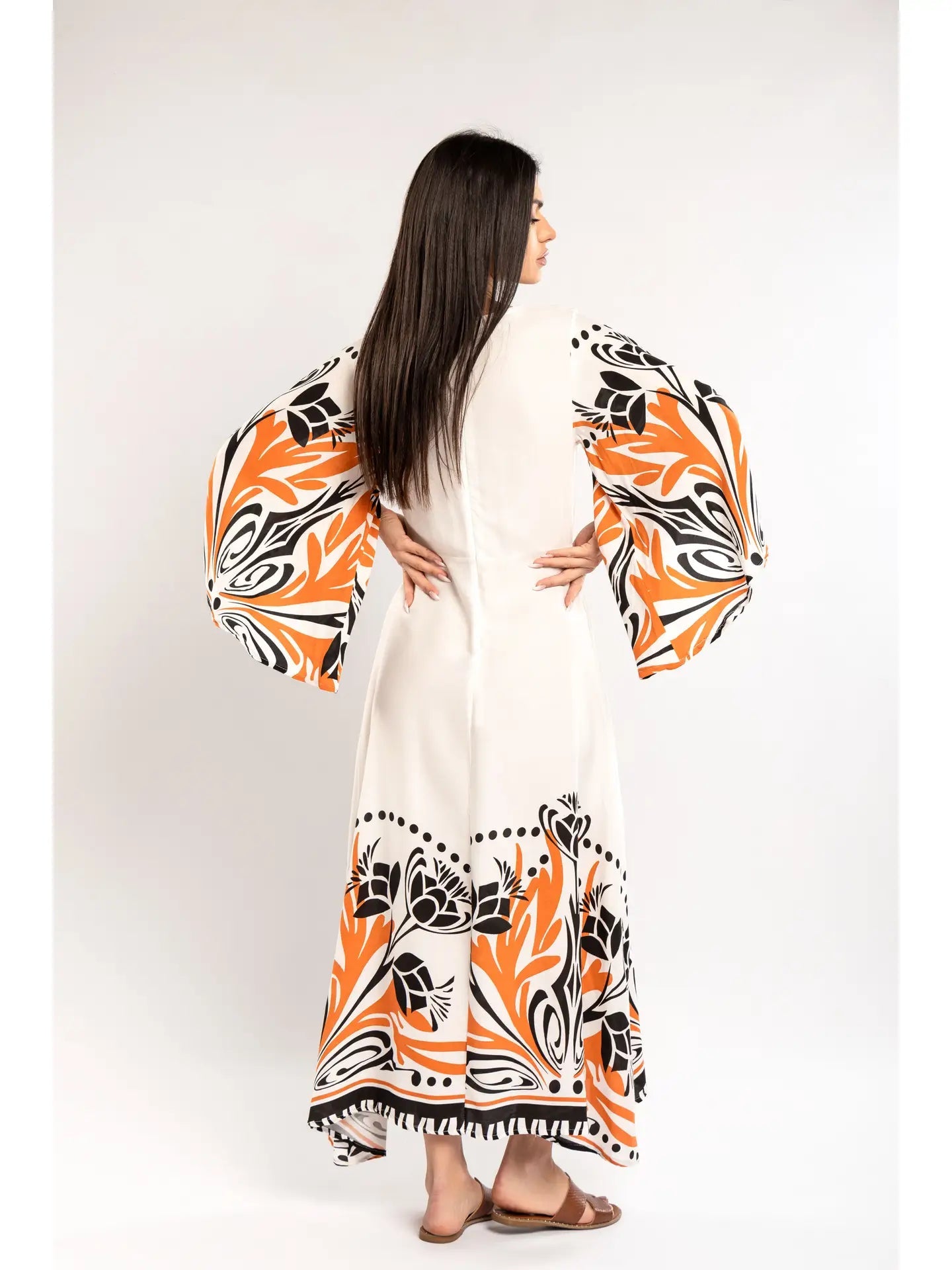 Woman wearing a long, patterned dress with orange and black designs on a white background - back view hands on hips