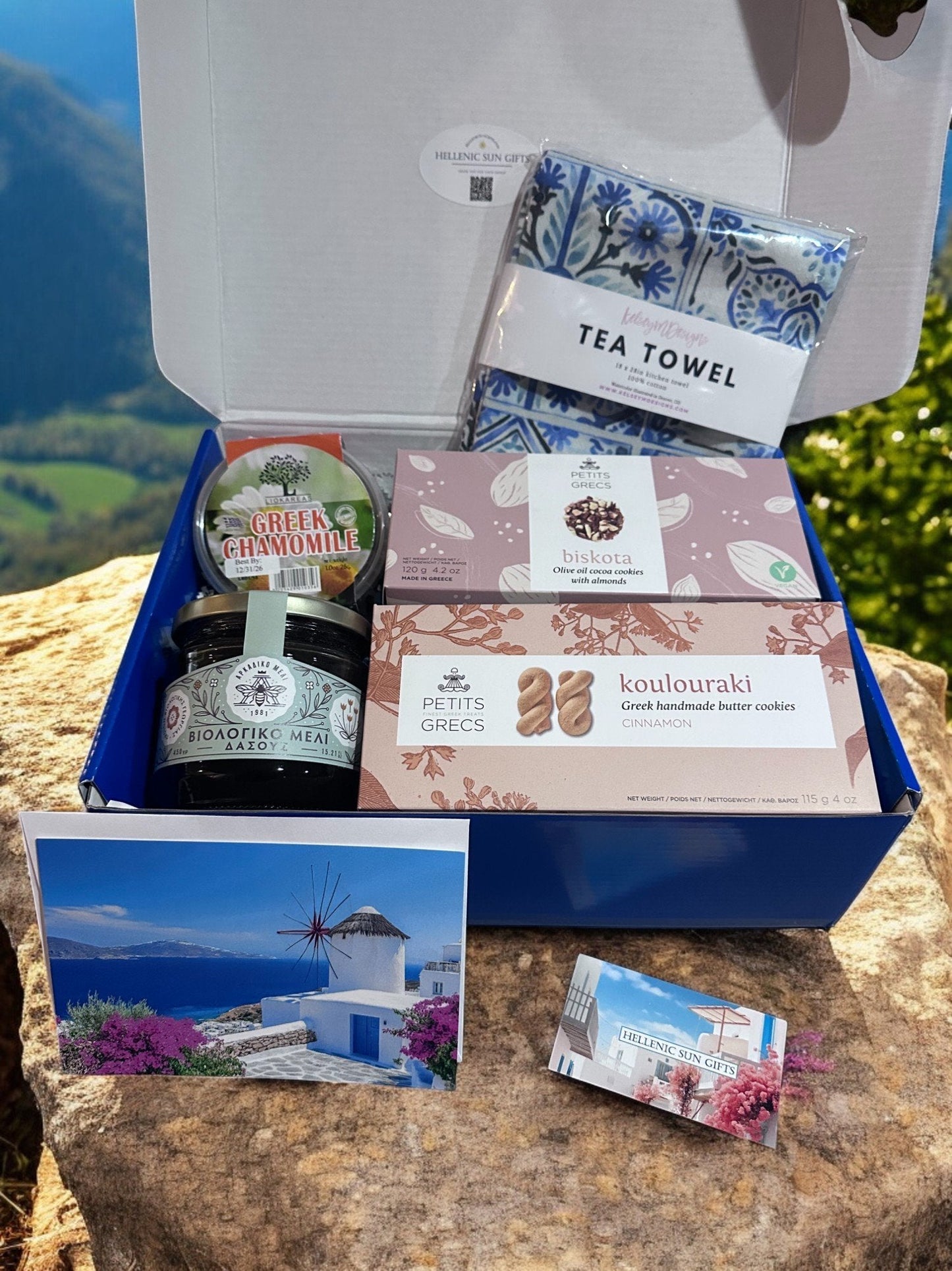 Gift box with Greek-themed products including a tea towel, cookies, and a jar of honey on a stone surface with a scenic background