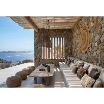 A luxury outdoor patio seating area with a stone wall, wooden ceiling, built-in cushioned sofa, woven poufs, and a central wooden table, overlooking a blue sea and sky.