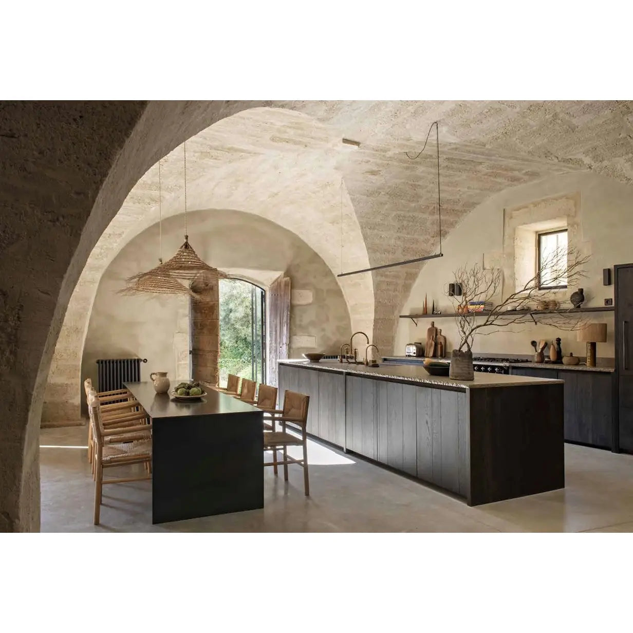 A modern kitchen and dining area set within a rustic stone building featuring arched ceilings, a dark wood island, a dining table with wooden chairs, and pendant lighting.