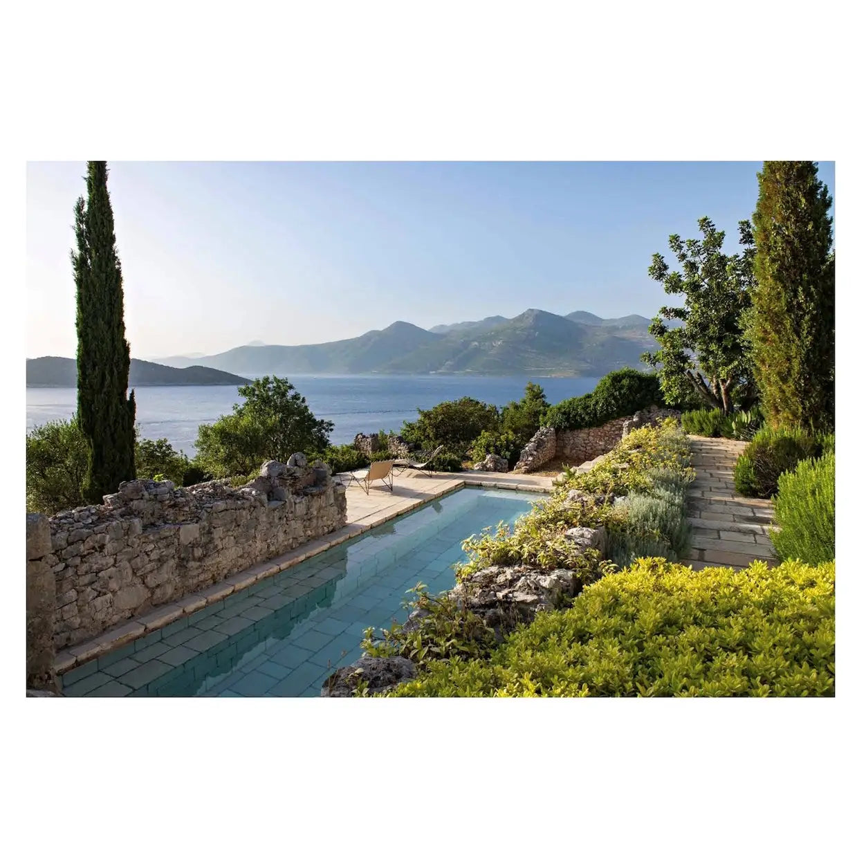 A scenic outdoor landscape with a long tiled swimming pool running through a landscaped garden, stone walls, tall cypress trees, and a distant sea and mountain view under a clear blue sky.