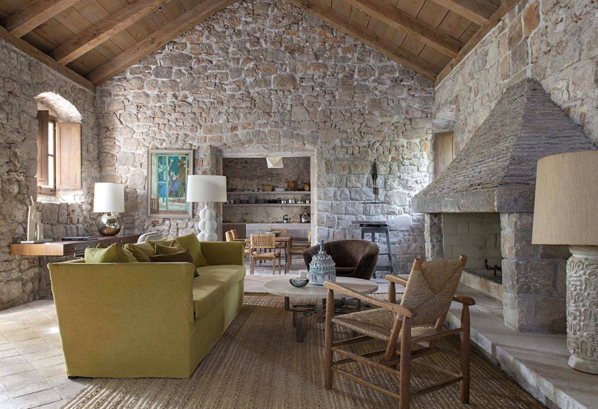 A rustic stone-walled living room with a large olive green sofa, a woven armchair, a low coffee table, and a massive stone fireplace under a wooden-beamed vaulted ceiling.