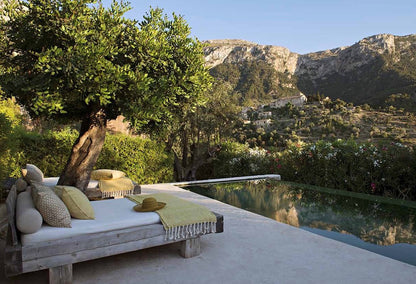 A luxury outdoor daybed with white cushions and yellow-green throw pillows sits beside a modern infinity pool, set under a large olive tree with a mountain village and rocky cliffs in the background.