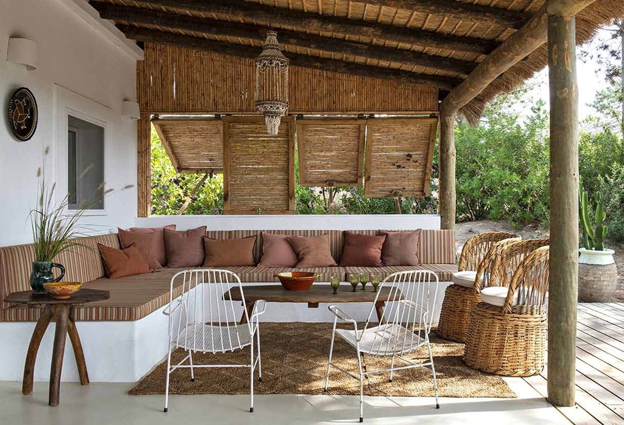 A rustic outdoor patio seating area with a long built-in bench, brown and rust cushions, white wire chairs, a small wooden side table, woven wicker chairs, a thatched roof, and greenery in the background.