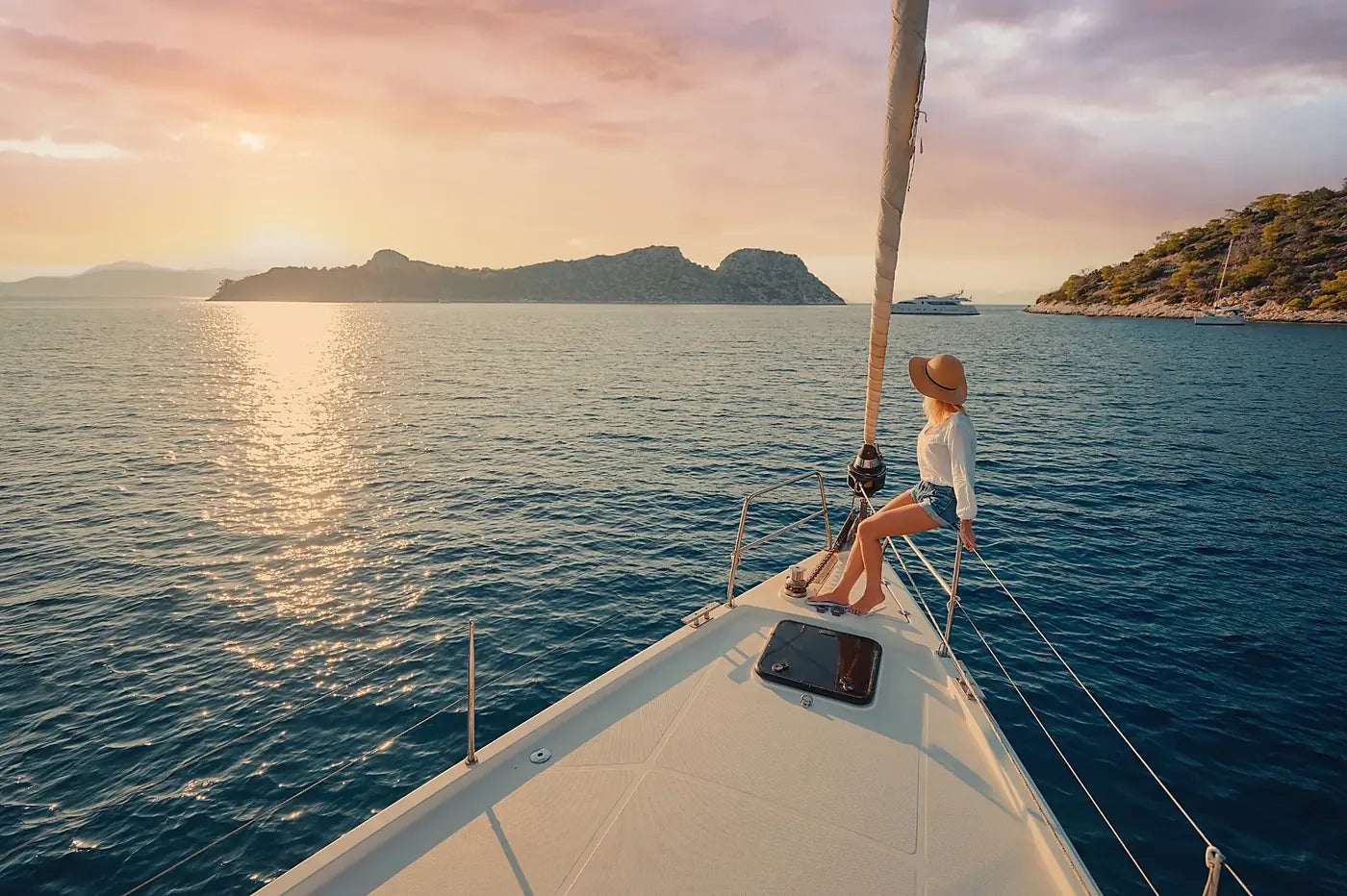 Woman sitting on a sailboat's bow with a scenic view of mountains and sunset