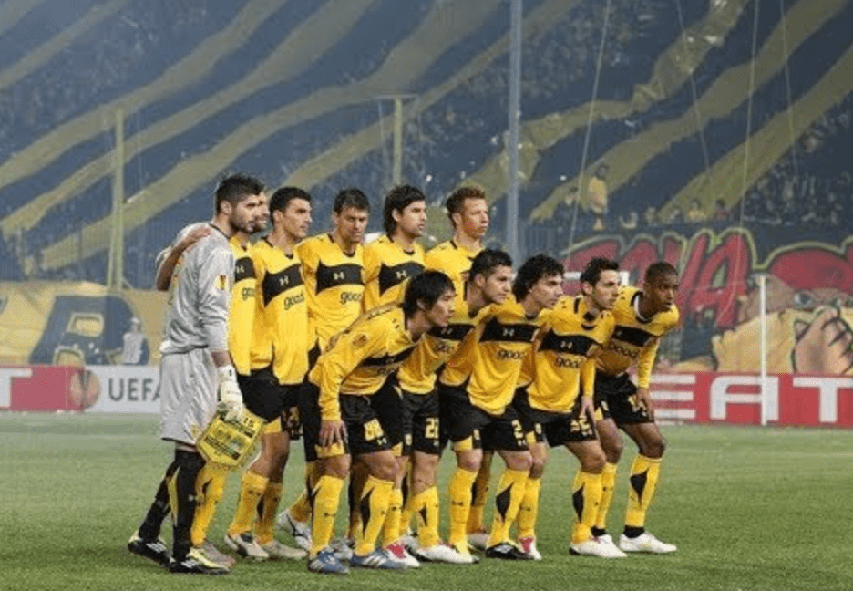 Greek Aris soccer players in yellow and black uniforms standing on a field with a blurred stadium background
