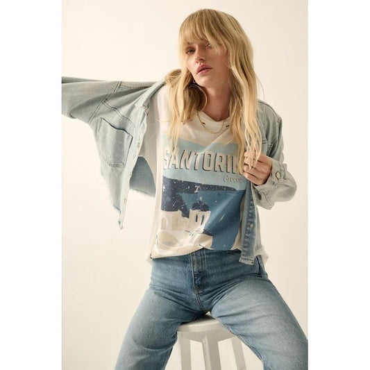 Front of blond woman wearing a distressed graphic tee with "Santorini Greece" and scenes of the domed churches and sea, along with jeans and a denim jacket, and sitting on a stool 