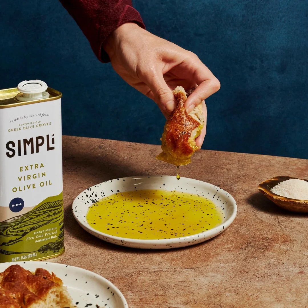 Person dipping bread into a dish of olive oil with a can of Simpli Extra Virgin Olive Oil in the background