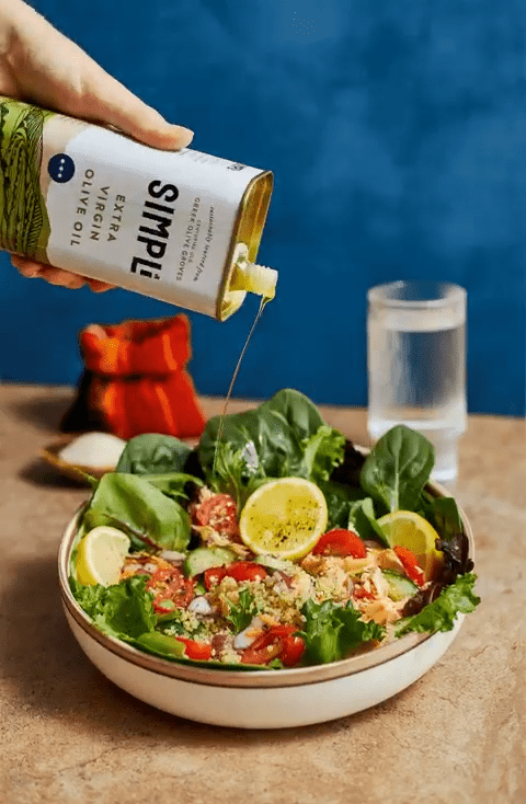 Person pouring Simple brand olive oil over a salad on a wooden table with a blue background