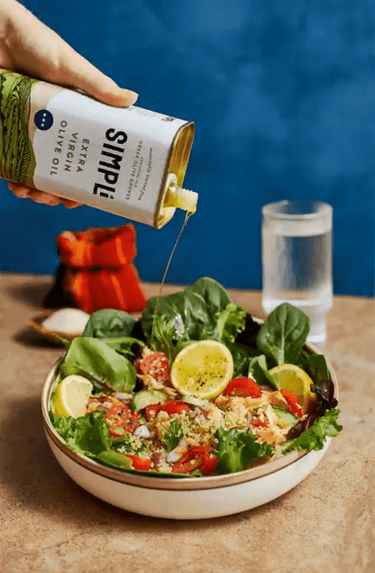 Person pouring Simple brand olive oil over a salad on a wooden table with a blue background